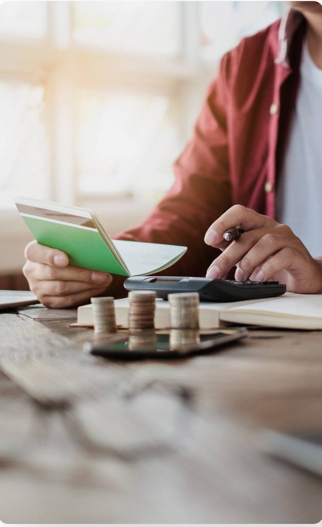 A person at a desk using a calculator with stacked coins in the foreground and holding an open book in one hand.