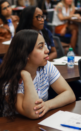 A person with long hair sits at a table, attentively listening, with a notepad and pen in front. Other attendees are visible in the background.