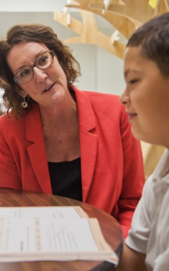 A woman in a red blazer talks with a boy who is looking at a sheet of paper on a table.