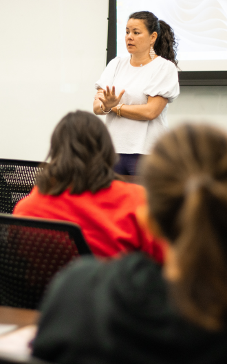 A woman in a white shirt is speaking in front of a classroom, gesturing with her hands. Students are seated, facing her.